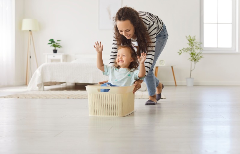 Young happy mother having fun at home together with child girl riding her in toy basket